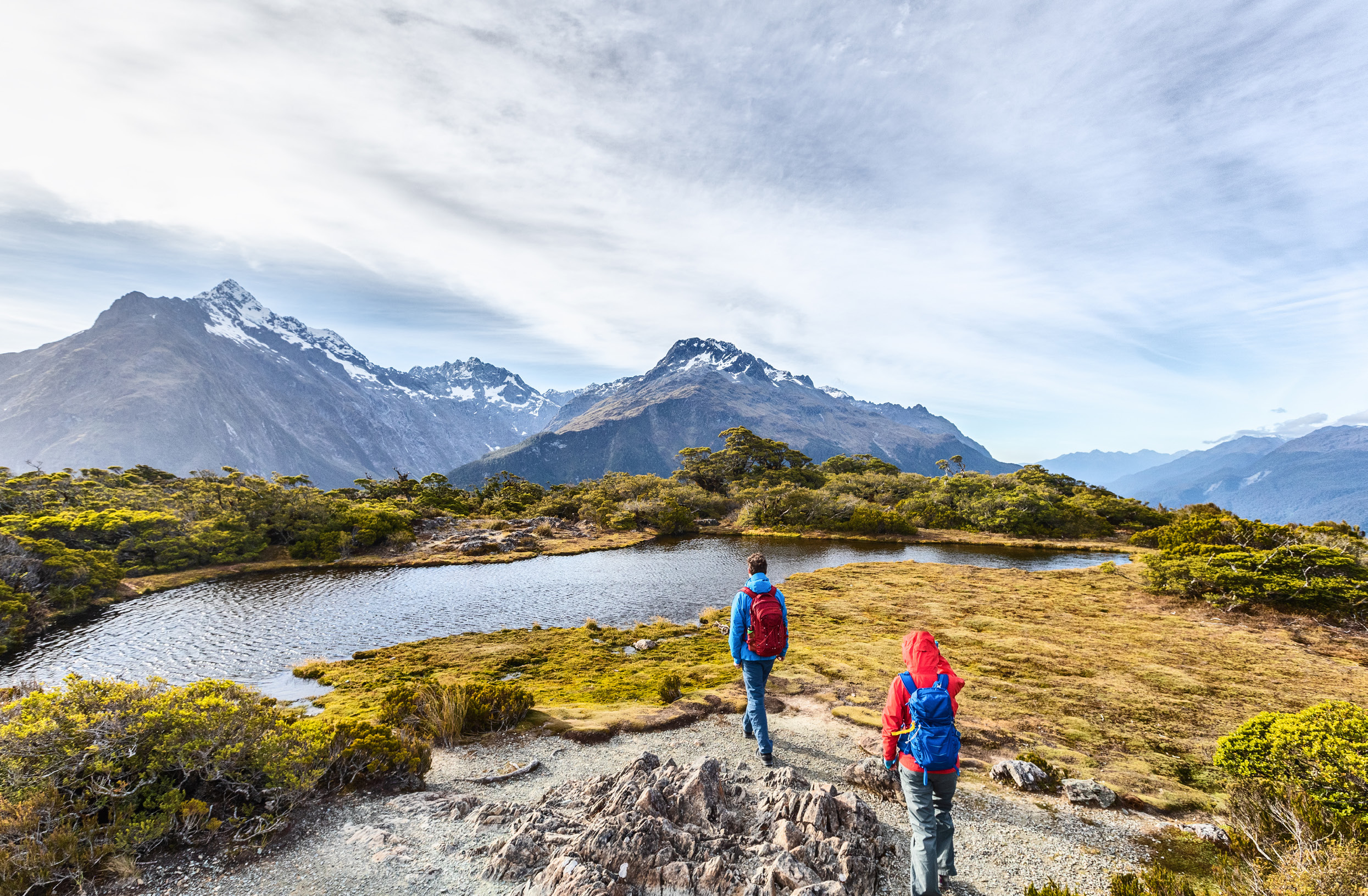 Two hikers walking beside an alpine lake surrounded by mountains