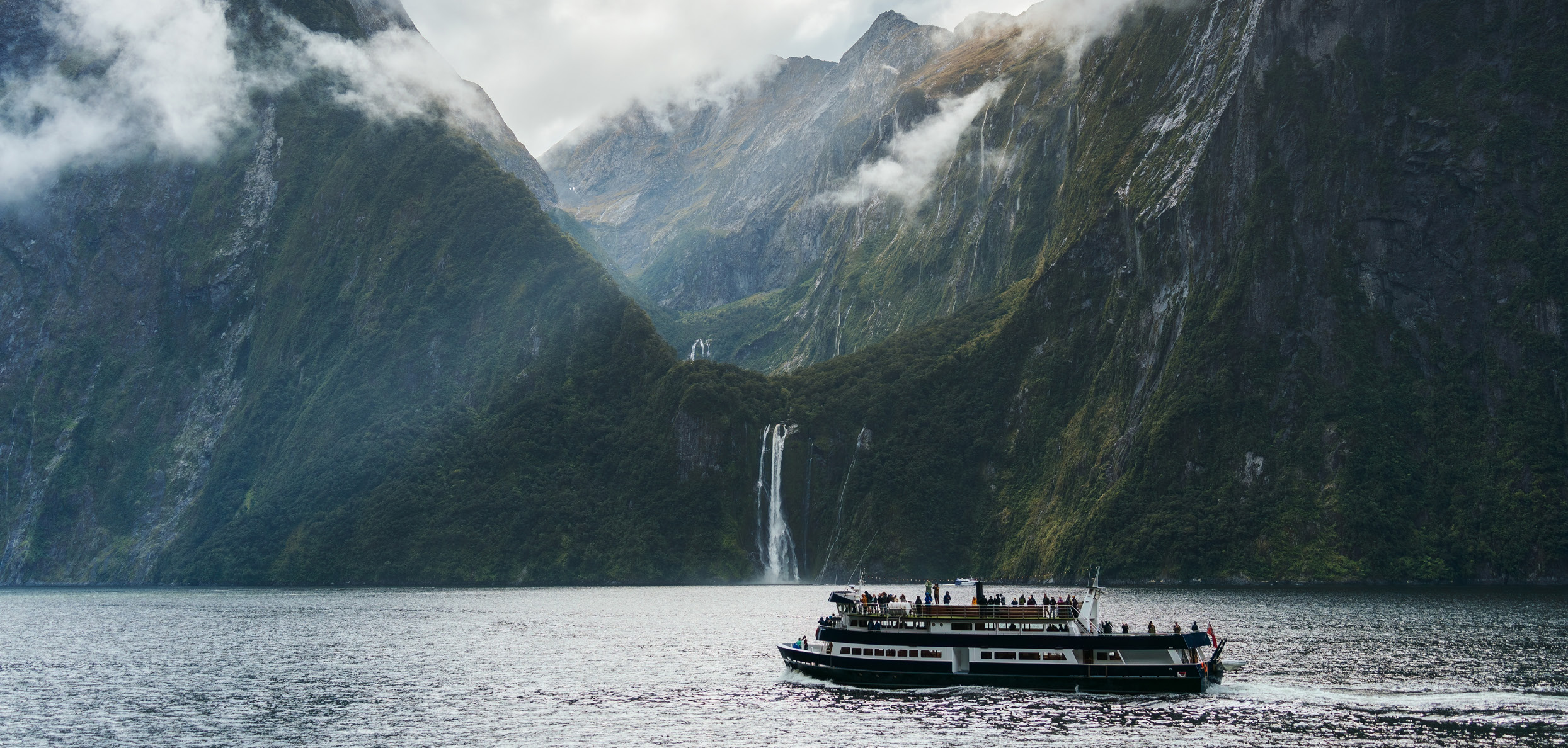 A cruise boat passing waterfalls and steep forested cliffs in Fiordland